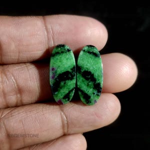 Op de afbeelding: Twee ovale edelstenen met een levendige groene basiskleur, geaccentueerd door zwarte en rode insluitsels. De stenen worden in een hand gehouden, waardoor hun gepolijste oppervlakken en natuurlijke patronen worden getoond. De edelstenen zijn waarschijnlijk voor het maken van sieraden.