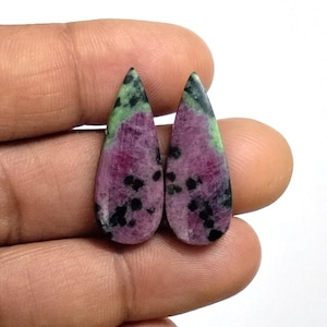 Può includere: Due cabochon a forma di goccia con un motivo maculato di magenta intenso, verde vibrante e punti neri sparsi. Queste pietre levigate presentano una superficie liscia e arrotondata con una punta definita e inclusioni minerali naturali.