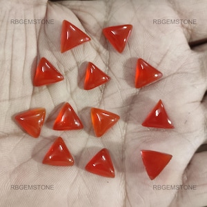 Può includere: Una collezione di gemme triangolari lucide in una vibrante tonalità arancione. Le gemme sono disposte su una superficie chiara, con il testo "RBGEMSTONE" visibile negli angoli. Queste gemme sono ideali per la creazione di gioielli o collezionismo.