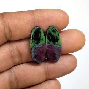Può includere: Due cabochon di forma ovale caratterizzati da una distinta combinazione di colori composta da magenta intenso, verde vibrante e inclusioni nere opache. Queste pietre preziose levigate presentano una superficie liscia e riflettente.