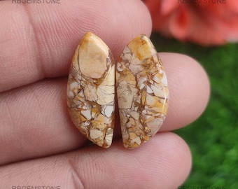 May include: Two polished, teardrop-shaped gemstones with a mosaic pattern of tan, cream, and brown. The stones are held between fingers, with the text "RBGEMSTONE" visible in the corners.