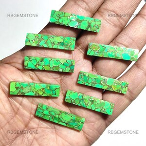 May include: Eight green and gold turquoise cabochons, cut into rectangular shapes, are displayed on a hand. The cabochons are all different sizes and have a variety of green and gold patterns.
