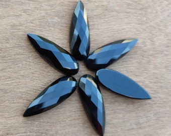 May include: Seven black faceted teardrop-shaped gemstone beads. The beads are arranged in a starburst pattern on a wooden surface.