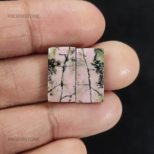 Può includere: Due pietre preziose rettangolari con una base rosa e venature nere. Le pietre preziose hanno macchie verdi e nere. Il testo "RBGEMSTONE" è visibile nell'immagine.