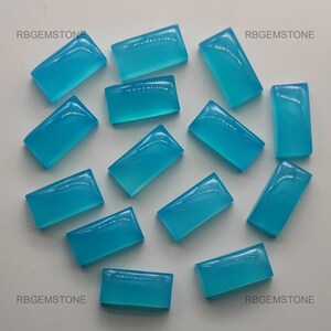 Op de afbeelding: Een verzameling van 14 turquoise blauwe rechthoekige cabochon edelstenen. De edelstenen zijn glad en gepolijst, met een lichtblauwe tint. De edelstenen zijn gerangschikt op een wit oppervlak.