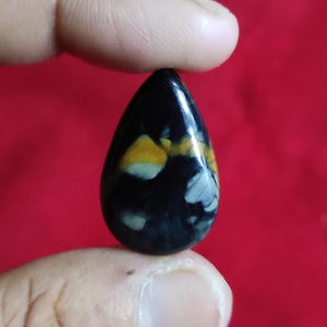 Puede incluir: Una piedra preciosa negra en forma de l&aacute;grima con remolinos blancos y amarillos. La piedra preciosa est&aacute; siendo sostenida por un dedo.
