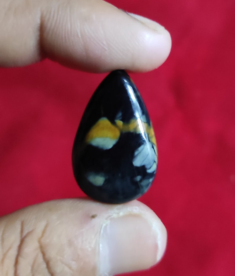 Puede incluir: Una piedra preciosa negra en forma de l&aacute;grima con remolinos blancos y amarillos. La piedra preciosa est&aacute; siendo sostenida por un dedo.