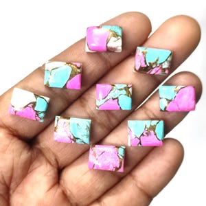 Puede incluir: Nueve gemas cuadradas en una mano, mostrando una mezcla de rosa, turquesa, blanco y dorado. Las gemas tienen patrones y texturas únicos. Adecuadas para la fabricación de joyas. Cada piedra mide aproximadamente 1 cm x 1 cm.