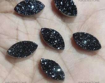 May include: Six black, oval-shaped, gemstone cabochons with a sparkly, textured surface.  The cabochons are arranged in a semi-circle on a dark surface.  RBGemstone is printed on the surface.