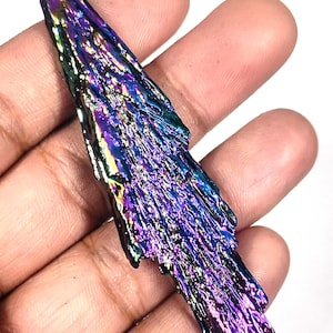 Puede incluir: Un cristal de cianita iridiscente con una superficie texturizada. La piedra muestra un espectro de colores, incluyendo púrpura, azul y verde. El cristal se sostiene en una mano, mostrando su forma natural y variaciones de color únicas.
