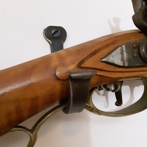 May include: A close-up of a wooden-stocked antique firearm with a brass trigger guard and a metal hook on the side. The firearm is mounted on a wall with a metal hook.