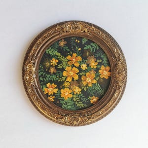May include: A round, vintage-style painting featuring yellow flowers with green leaves against a dark background. The artwork is framed in an ornate, gold-colored oval frame, adding a touch of classic elegance.