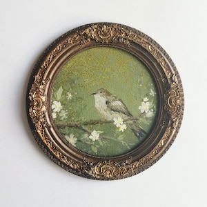 May include: A round framed painting featuring a bird perched on a branch with white flowers. The bird has gray and brown feathers, set against a green background. The ornate frame is gold-colored.