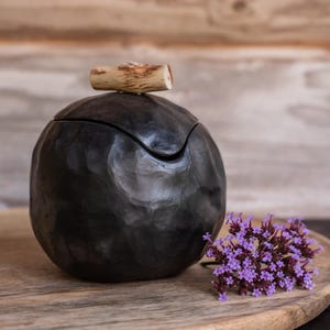 Op de afbeelding: Een ronde, donker antracietkleurige houder met een getextureerd, gehamerd oppervlak en een deksel met een kleine handgreep van een lichtgekleurde houten tak. Een trosje kleine, felpaarse bloemen ligt naast de houder.