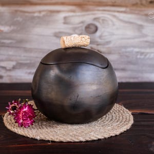 May include: A dark, spherical container with a lid, resting on a woven coaster. A cork sits atop the lid. A cluster of pink flowers is to the left. The background is a wooden surface.