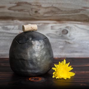 May include: A round, dark gray ceramic jar with a hammered, textured surface and a matching lid. A small, cylindrical light-colored wooden knob sits atop the lid. A bright yellow chrysanthemum flower is placed next to the jar.