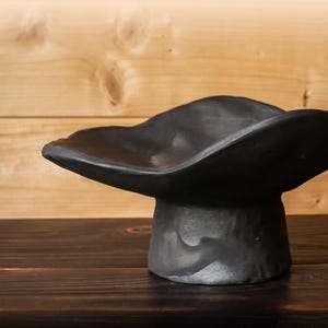 May include: A handcrafted, black ceramic bowl with a wide, undulating rim. The bowl sits atop a short, textured pedestal base. The dark color and organic shape suggest a modern, minimalist aesthetic. The bowl is displayed on a dark wooden surface.