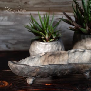 Op de afbeelding: Een handgemaakte, grijs getinte, langwerpige plantenbak met drie kleine voetjes, tentoongesteld op een donker houten oppervlak. Twee extra getextureerde plantenbakken met groene vetplanten staan op de achtergrond. De tekst "Clay & Wood Design" is zichtbaar.