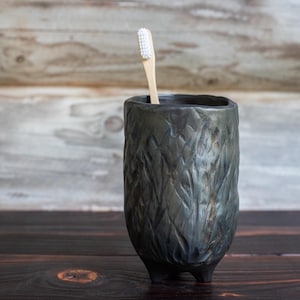 Könnte beinhalten: Ein dunkelgrauer Keramik-Zahnbürstenhalter mit einem strukturierten, blattartigen Muster. Eine Holzzahnbürste mit weißen Borsten ist darin platziert. Der Halter steht auf einer dunklen Holzoberfläche.