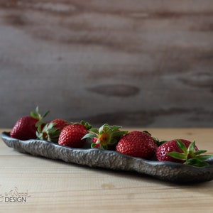 Op de afbeelding: Een lange, zwarte, getextureerde keramische serveerschaal met een rij rode aardbeien. De schaal staat op een licht houten oppervlak.