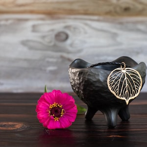 May include: A black, three-legged ceramic bowl with a hammered texture containing a black cord necklace with a gold-toned, openwork leaf-shaped pendant. A vibrant pink zinnia flower with a yellow center is positioned next to the bowl.