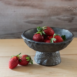 Può includere: Una ciotola a piedistallo grigio scuro e strutturata piena di fragole rosse fresche con foglie verdi. Due fragole poggiano sulla superficie di legno davanti alla ciotola. La ciotola ha un bordo ruvido e organico e poggia su una base strutturata.