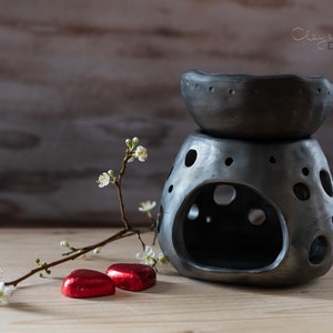 Può includere: Un diffusore di oli essenziali in ceramica grigia con una parte superiore a forma di ciotola e una base perforata. Il diffusore è su una superficie di legno, con un rametto di fiori bianchi e due caramelle rosse a forma di cuore. Design di Clay & Wood Design.