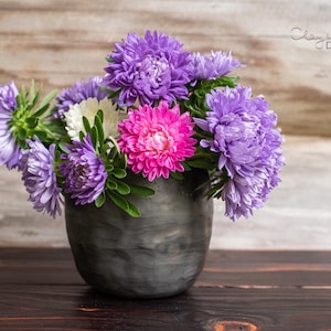 Op de afbeelding: Een donkergrijze vaas gevuld met een levendig boeket van paarse, roze en witte asters. De bloemen staan in volle bloei, met weelderig groen blad. De vaas staat op een donker houten oppervlak.