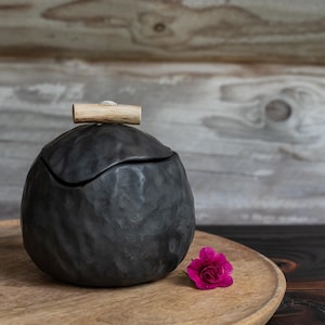 Puede incluir: Un tarro de cerámica negro mate de forma redonda con una superficie texturizada e irregular y una tapa a juego. La tapa presenta un pequeño mango cilíndrico de madera clara en el centro. Una flor magenta brillante descansa junto a la base.