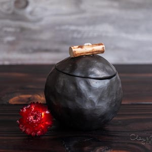 Könnte beinhalten: Eine schwarze Keramikdose mit einer rauen, strukturierten Oberfläche. Die Dose hat einen Holzdeckel mit natürlicher Oberfläche. Die Dose steht auf einer dunklen Holzoberfläche.