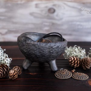 May include: A black ceramic bowl with three legs sits on a wooden surface. The bowl has a rough, textured surface and is filled with a necklace. Two round, bronze earrings with a textured design are in front of the bowl. The bowl is surrounded by small, white, fluffy plants.