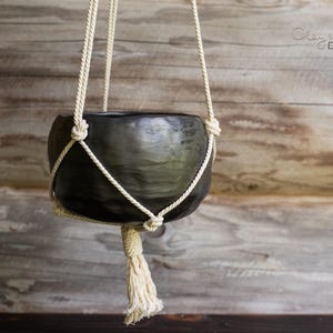 May include: A hanging planter with a dark gray, textured bowl suspended by cream-colored macrame rope. The rope is knotted to form a cradle for the bowl and a tassel hangs below. The background is a wooden wall.