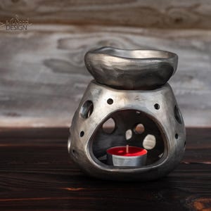 Può includere: Un diffusore di oli essenziali color argento con una parte superiore a forma di ciotola e una base con fori decorativi. Una candela rossa accesa si trova all'interno della base. Il design è rustico e fatto a mano.