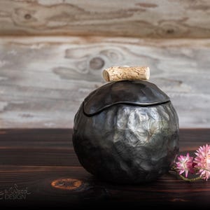 May include: Black ceramic jar with a wooden lid, sitting on a dark wood surface with a pink flower in the foreground. The jar has a textured surface and a unique shape.  Clay & Wood Design.