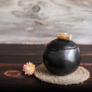 Könnte beinhalten: Ein schwarzer Keramiktopf mit Holzdeckel, auf einem gewebten Untersetzer. Eine getrocknete Blume mit rosa und gelben Blütenblättern liegt daneben. Der Topf ist rund und hat eine glatte, dunkle Oberfläche.