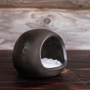 Può includere: Un letto per animali domestici in ceramica arrotondata grigio scuro con una grande apertura. L'interno è riempito di materiale bianco, possibilmente biancheria da letto. Il letto è su una superficie di legno scuro, con uno sfondo di legno chiaro.