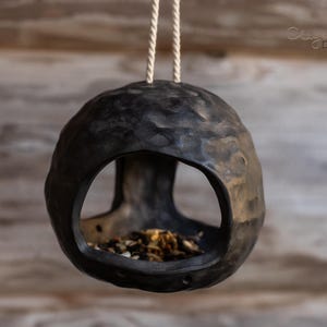 Peut inclure: Un nichoir gris foncé texturé, avec une grande ouverture et une forme ronde, suspendu par une corde naturelle. La mangeoire est remplie de graines pour oiseaux et a un aspect rustique et fait à la main. L'arrière-plan est une surface en bois floue.