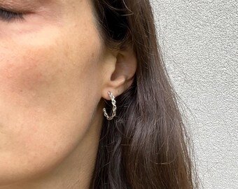 May include: Silver hoop earrings with a braided design. The earrings are worn in a person's ear.