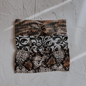 May include: Three fabric headbands with a twisted knot detail. The top headband features a brown and black plaid pattern. The middle headband has a black and white yin-yang symbol print. The bottom headband displays a brown and black snakeskin pattern.