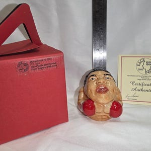 May include: A ceramic figurine of a boxer with red boxing gloves. The figurine is tan with black hair and facial features. A red box with a handle and text is on the left. A certificate of authenticity is on the right.