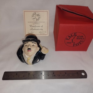 May include: A ceramic figurine of Charlie Chaplin in a black suit and bowler hat, with a painted face and mustache. The figurine is next to a red box with the words "FACE FRANCIS POTS" and a certificate of authenticity. A metal ruler is also present.