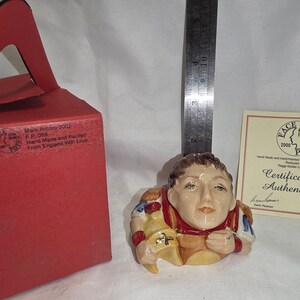 May include: A ceramic Mark Antony character jug by Face Pots, measuring approximately 3.5 inches in height. The figure features a tan face, red and gold armor, and is accompanied by a red presentation box and a certificate of authenticity.
