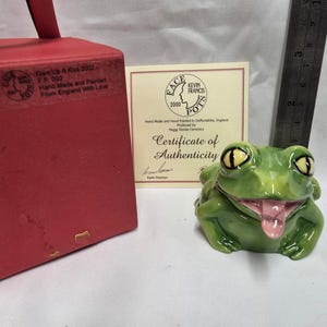 May include: A green ceramic frog figurine with yellow eyes and a pink tongue, accompanied by a red box, a certificate of authenticity, and a metal ruler showing a height of approximately 2.5 inches. The box features text including Give Us A Kiss 2002.