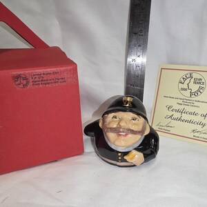 May include: A ceramic figurine of a police officer wearing a black helmet with VR insignia, accompanied by a red box, a certificate of authenticity, and a metal ruler showing a height of approximately 3.5 inches for the figurine.