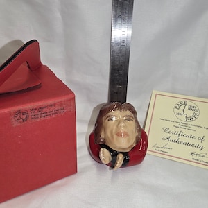 Pode incluir: Uma estatueta de cerâmica da cabeça de uma pessoa com cabelo castanho, usando um casaco vermelho e uma camisa preta. Uma caixa vermelha e um certificado de autenticidade estão ao lado da estatueta. Uma régua de metal está atrás da estatueta.