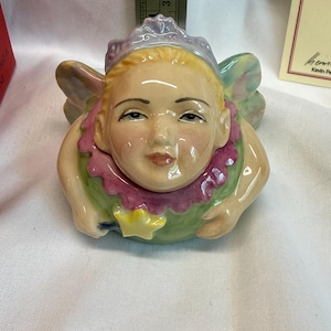 May include: A ceramic fairy figurine with blonde hair, a purple crown, and a pink ruffled collar. The figure holds a yellow star-tipped wand and has pastel-colored wings. A metal ruler behind the figure shows a height of approximately 2.5 inches.