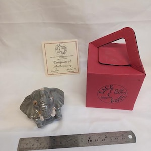May include: A grey ceramic elephant figurine with a detailed trunk and tusks. A red box with the text "FACE KEVIN FRANCIS POTS 2000" and a certificate of authenticity are also present. A metal ruler measures up to 15 cm.