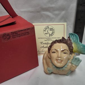 May include: A ceramic merman figurine with brown hair and a teal tail, accompanied by a red box and a certificate of authenticity. A metal ruler is visible, showing the figurine is approximately 3.5 inches tall. The box label reads Mygard The Merman 2001.
