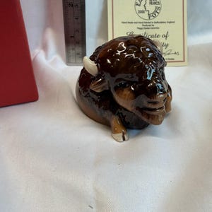 May include: A ceramic figurine of a bison, primarily brown with lighter brown and white accents. The bison is in a resting position. A ruler is next to the bison. A certificate is behind the bison.