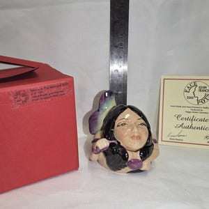 Może przedstawiać: Ręcznie malowana ceramiczna figurka syreny z czarnymi włosami i fioletowymi akcentami. Figurka jest dostarczana z certyfikatem autentyczności i czerwonym pudełkiem. Twarz syreny jest szczegółowa.